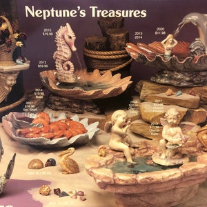 May include: A collection of decorative garden fountains featuring cherubs, seahorses, dolphins, and a pelican. The fountains are made of a light brown stone-like material and are decorated with seashells and other nautical elements. The fountains are displayed on a background of sand and rocks.