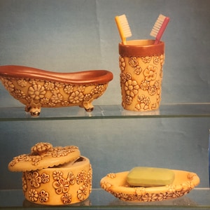 Puede incluir: Un juego de cuatro accesorios de baño con un patrón floral. El juego incluye un portacepillos de dientes, un jabonera, una pequeña bañera y un frasco con tapa. Los artículos son todos de cerámica y tienen un color marrón claro con un diseño floral.