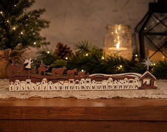 May include: A wooden advent calendar featuring a laser-cut nativity scene silhouette. The base displays numbers 1 through 24, with a row of buildings and figures including camels, animals, and an angel. A star sits atop a stable on the far right.