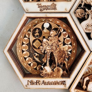 Könnte beinhalten: Ein sechseckiger Holzrahmen mit einem detaillierten Laser-Cut-Design, das einen Charakter aus dem Videospiel Nier: Automata zeigt. Der Rahmen ist mit "Nier: Automata" beschriftet und hat ein kreisförmiges Design mit einer stilisierten Figur in der Mitte. Der Rahmen ist Teil eines größeren Sets von sechseckigen Rahmen, die jeweils einen anderen Charakter oder ein anderes Design zeigen.