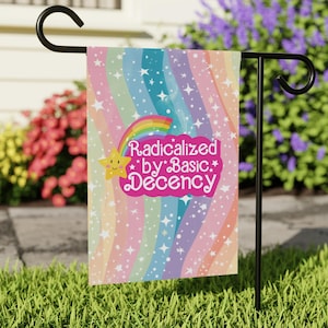 May include: A garden flag with a rainbow design and the text "Radicalized by Basic Decency." The flag features a rainbow, a smiling star, and white stars on a pastel background. The flag is displayed on a black metal stand in a grassy garden.