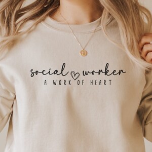 Peut inclure: Un sweat-shirt beige avec le texte "social worker" et un symbole de cœur, suivi du texte "a work of heart".