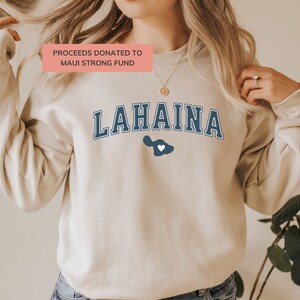 May include: A beige sweatshirt with the text "LAHAINA" in navy blue with a white heart inside a navy blue outline of the island of Maui. The text "PROCEEDS DONATED TO MAUI STRONG FUND" is in pink.