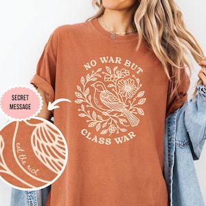 Puede incluir: Una camiseta de color óxido con un gráfico de color crema de un pájaro rodeado de enredaderas florales. El texto dice NO WAR BUT CLASS WAR, con un mensaje oculto más pequeño en el diseño que dice eat the rich en cursiva.