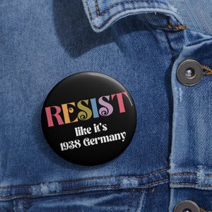May include: A black button with the word "RESIST" in colorful letters. Below the word is the phrase "like it's 1938 Germany" in white. The button is pinned to a denim jacket.