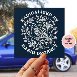 May include: A navy blue square magnet or sticker with white text and a floral bird design. The text reads "RADICALIZED BY BASIC DECENCY" in a circular pattern around the bird illustration. The product is held in front of a blue car.