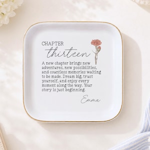 May include: A square white ceramic trinket dish with rounded corners and a gold-colored rim. The center features a printed illustration of a pink carnation and black text that reads: CHAPTER thirteen, A new chapter brings new adventures, and Emma.