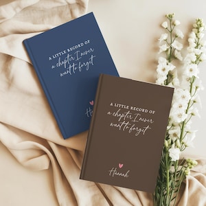 May include: Two hardcover journals, one navy blue and one dark brown, each featuring the white printed text A LITTLE RECORD OF a chapter I never want to forget. The brown journal includes a small pink heart and the name Hannah in white script.