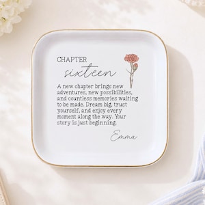 May include: A square white ceramic trinket dish with rounded corners and a gold-colored rim. The center features a floral illustration of a carnation and text that reads: CHAPTER sixteen, A new chapter brings new adventures, and the name Emma.