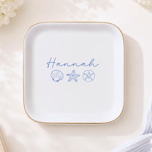 Puede incluir: Un plato cuadrado de cerámica blanca con esquinas redondeadas y un borde fino de color dorado. El centro muestra el nombre Hannah en letra cursiva azul, junto a ilustraciones de línea azul de una concha, una estrella y un dólar de arena.