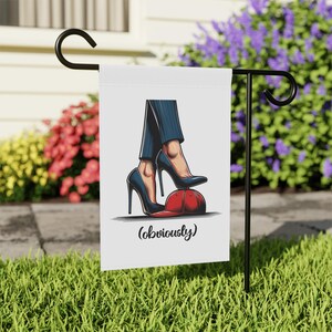 May include: A white garden flag with a cartoon illustration of a pair of high heels stepping on a red baseball cap. The text '(obviously)' is printed below the image.