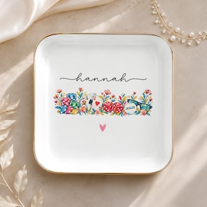May include: A square white ceramic trinket dish with a gold-toned rim. The center features the name hannah in black script above a colorful illustration of poker chips, playing cards, red dice, and a dealer button nestled in flowers.