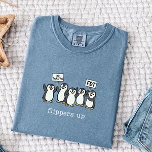 May include: A blue Comfort Colors t-shirt featuring a graphic of five cartoon penguins holding signs that read "NO TARIFFS!" and "FDT". The text "flippers up" is printed below the penguins.