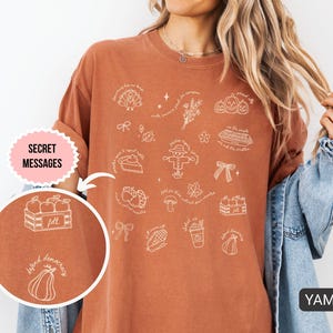 May include: Burnt orange t-shirt featuring white line art illustrations of autumn-themed items, such as pumpkins, a scarecrow, and a pie. The shirt has the words "Secret Messages" and "YAM" printed on it.