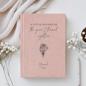 Peut inclure: Un journal à couverture rigide de couleur rose poudré avec une finition en tissu texturé. La couverture présente un texte noir indiquant A LITTLE RECORD OF the year I turned eighteen, un dessin au trait d'un bouquet, le nom Hannah et 2026.