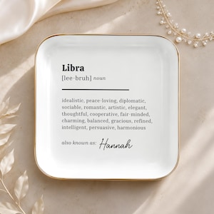 May include: A square white ceramic trinket tray with rounded corners and a gold-toned rim. The center features black text defining Libra with adjectives like idealistic, peace-loving, and diplomatic, followed by the custom text also known as: Hannah.