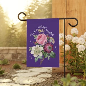 May include: A purple garden flag with a floral bouquet design and the words "practice radical empathy" in white script. The bouquet features pink, white, and burgundy flowers with green leaves. The flag is displayed on a black metal stand.