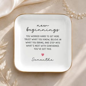 Puede incluir: Un plato cuadrado de cerámica blanca con esquinas redondeadas y un borde dorado. El centro presenta texto negro que dice: new beginnings, YOU WORKED HARD TO GET HERE. TRUST WHAT YOU KNOW, BELIEVE IN WHAT YOU BRING, AND STEP INTO WHAT'S NEXT WITH CONFIDENCE. YOU'VE GOT THIS. Un pequeño corazón rosa y el nombre Samantha están impresos debajo.