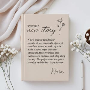 May include: A beige hardcover journal featuring the black printed text "WRITING A new story" and a floral line drawing. Below, a paragraph reads: "A new chapter brings new opportunities, new challenges, and countless memories waiting to be made."