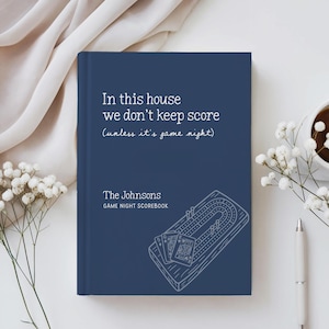 May include: A dark blue hardcover scorebook with white text that reads In this house we don't keep score (unless it's game night), The Johnsons, and GAME NIGHT SCOREBOOK, featuring a line drawing of a cribbage board and playing cards.