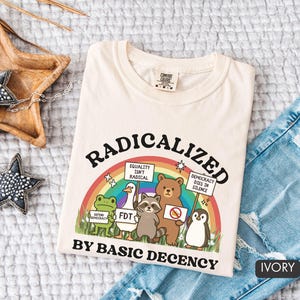 May include: Ivory-colored t-shirt with the text "RADICALIZED BY BASIC DECENCY" above a rainbow and cartoon animals holding signs. The shirt is folded on top of a pair of blue jeans.