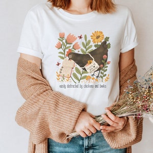 May include: White t-shirt with a black chicken illustration holding a book that says "Gluck 4 you Reading". The illustration is surrounded by flowers and mushrooms. The text "easily distracted by chickens and books" is printed below the illustration.