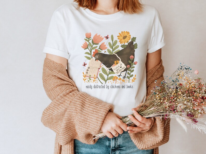 May include: White t-shirt with a black chicken illustration holding a book that says "Gluck 4 you Reading". The illustration is surrounded by flowers and mushrooms. The text "easily distracted by chickens and books" is printed below the illustration.
