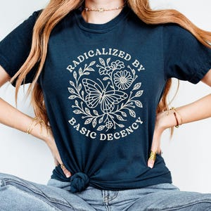 May include: Navy blue t-shirt featuring a white floral and butterfly design with the text "RADICALIZED BY BASIC DECENCY" in a circular arrangement. The shirt is knotted at the waist.