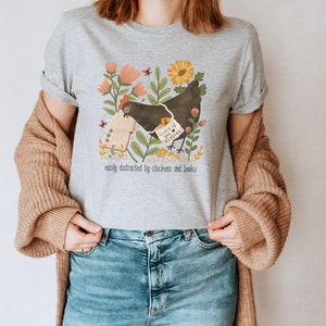 May include: Gray t-shirt with a black chicken reading a book. The book says "Such a Luck if You're Reading". The shirt has a floral design and the text "easily distracted by chickens and books".