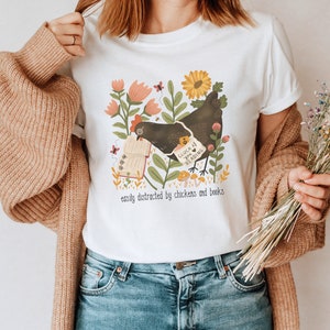 May include: White t-shirt with a graphic of a black chicken holding a book with the text "Cluck if you're reading" and the text "easily distracted by chickens and books".
