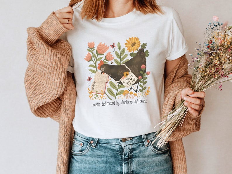 May include: White t-shirt with a graphic of a black chicken holding a book with the text "Cluck if you're reading" and the text "easily distracted by chickens and books".