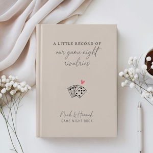 May include: A beige hardcover book titled A LITTLE RECORD OF our game night rivalries with a small illustration of playing cards and a pink heart, featuring the names Noah & Hannah and the text GAME NIGHT BOOK on the front cover.