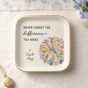 May include: A square white ceramic trinket dish with a gold-toned rim. The center features a colorful pom-pom graphic, a small pink heart, and the text: NEVER FORGET THE difference YOU MAKE, Coach Amy. The design is printed in black and script fonts.