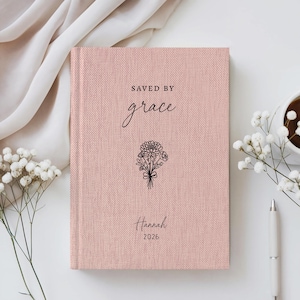 May include: A dusty pink hardcover journal with a textured fabric finish. The cover features the black printed text "SAVED BY grace" above a line-art illustration of a flower bouquet, with the name "Hannah" and the year "2026" printed below.