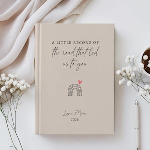 May include: A beige hardback book featuring the black text A LITTLE RECORD OF the road that led us to you, a small black line-art rainbow with a pink heart, and the text Love, Mom 2026 printed on the front cover in a clean, centred layout.