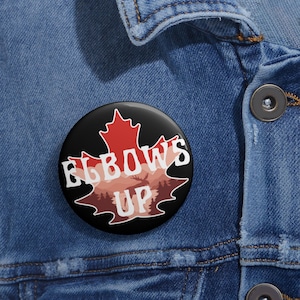 May include: A black button with a red maple leaf design. The leaf has a white outline and the words "Elbows Up" are printed in white on the leaf.
