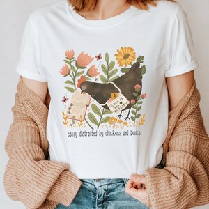 May include: White t-shirt with a black chicken holding a book that says "Cluck at you reading". The chicken is surrounded by flowers and mushrooms. The text below the image says "easily distracted by chickens and books".