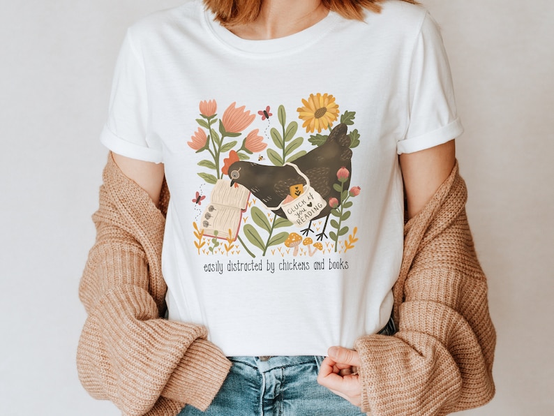 May include: White t-shirt with a black chicken holding a book that says "Cluck at you reading". The chicken is surrounded by flowers and mushrooms. The text below the image says "easily distracted by chickens and books".