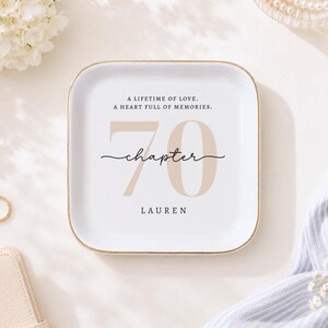 May include: A square white ceramic trinket dish with a gold-toned rim. The center features the text A LIFETIME OF LOVE. A HEART FULL OF MEMORIES. above a large beige number 70, the word chapter in black script, and the name LAUREN below.
