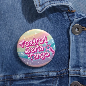 Może przedstawiać: Guzik w kolorze tęczy z białą gwiazdą i napisem "Foxtrot Delta Tango" w różowych literach.