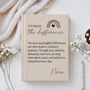 May include: A beige hardcover journal featuring a black line-art rainbow with a heart and the text: YOU MAKE the difference. The most meaningful differences are often made in ordinary moments. Through your patience, dedication, and care, you help others grow, learn, and believe in themselves every day. Nora.
