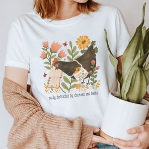 May include: White t-shirt with a black chicken reading a book surrounded by flowers and mushrooms. The text on the shirt reads "Cluck It, I'm Reading" and "easily distracted by chickens and books".
