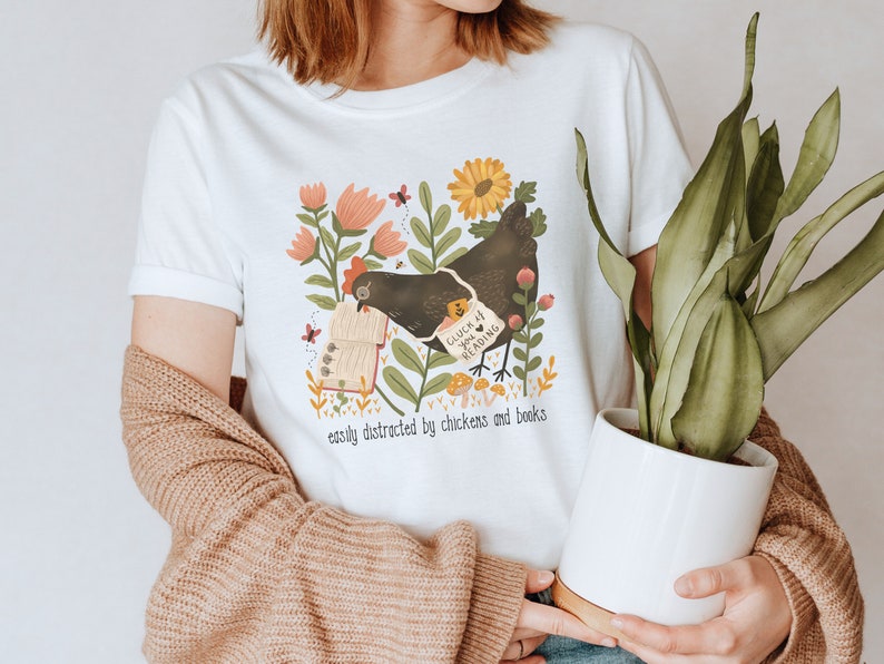 May include: White t-shirt with a black chicken reading a book surrounded by flowers and mushrooms. The text on the shirt reads "Cluck It, I'm Reading" and "easily distracted by chickens and books".