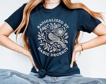May include: A dark navy blue short-sleeve t-shirt featuring a white line-art graphic of a bird perched among flowers and leaves. The text RADICALIZED BY BASIC DECENCY is printed in a circular arrangement around the central bird and floral design.
