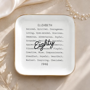 May include: A square white ceramic trinket dish with a gold-toned rim. The center features the name ELIZABETH, the word Eighty in black script, the year 1946, and a list of descriptive words including Beloved, Spirited, Courageous, and others.