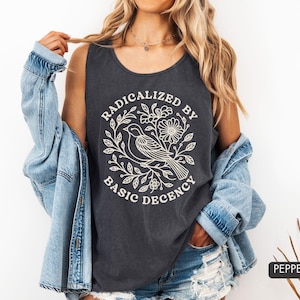 May include: A charcoal gray sleeveless tank top featuring a white line art graphic of a bird perched among flowers and leaves. The text RADICALIZED BY BASIC DECENCY is printed in a circular arrangement around the central bird and floral design.