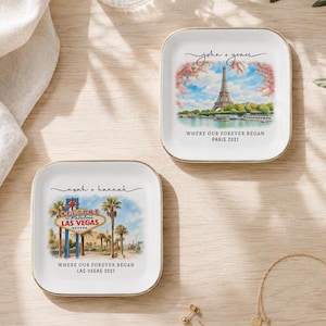 Puede incluir: Dos platos cuadrados de cerámica blanca para joyas con bordes dorados. Uno muestra la Torre Eiffel, el texto "john + grace" y "WHERE OUR FOREVER BEGAN PARIS 2027". El otro muestra el letrero de Las Vegas, "noah + hannah" y "WHERE OUR FOREVER BEGAN LAS VEGAS 2027".