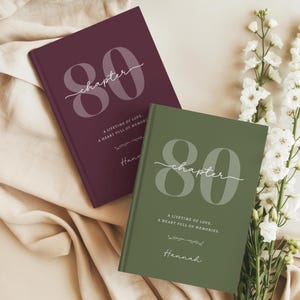 Peut inclure: Deux livres à couverture rigide avec des finitions mates en bordeaux profond et vert olive. Chaque couverture présente le grand nombre 80, le mot chapter en écriture cursive, et le texte A LIFETIME OF LOVE, A HEART FULL OF MEMORIES, avec le nom Hannah.