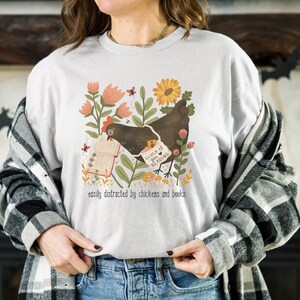 May include: A white t-shirt with a graphic of a brown chicken reading a book in a garden setting. The text on the shirt reads "Catch me if you can... reading" and "easily distracted by chickens and books".