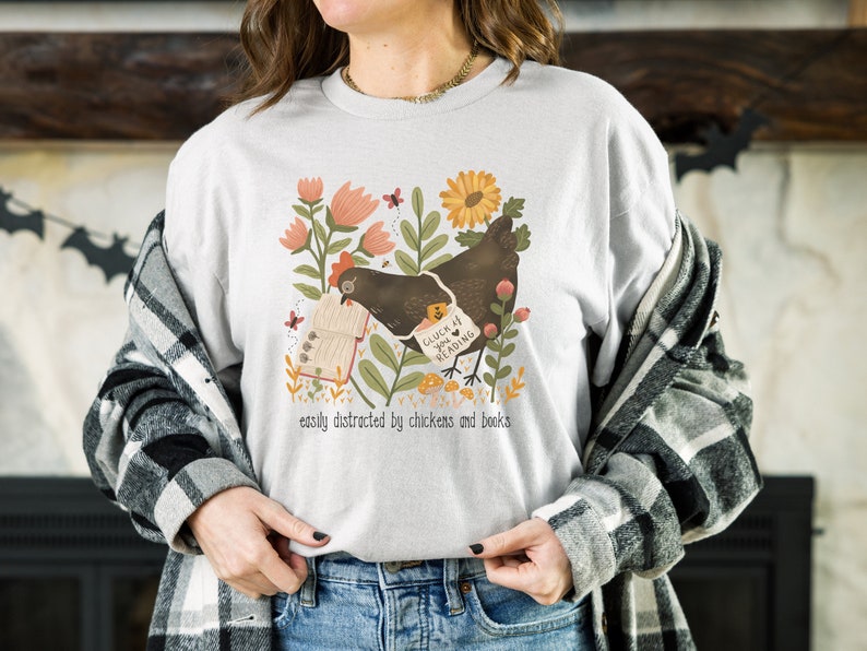 May include: A white t-shirt with a graphic of a brown chicken reading a book in a garden setting. The text on the shirt reads "Catch me if you can... reading" and "easily distracted by chickens and books".